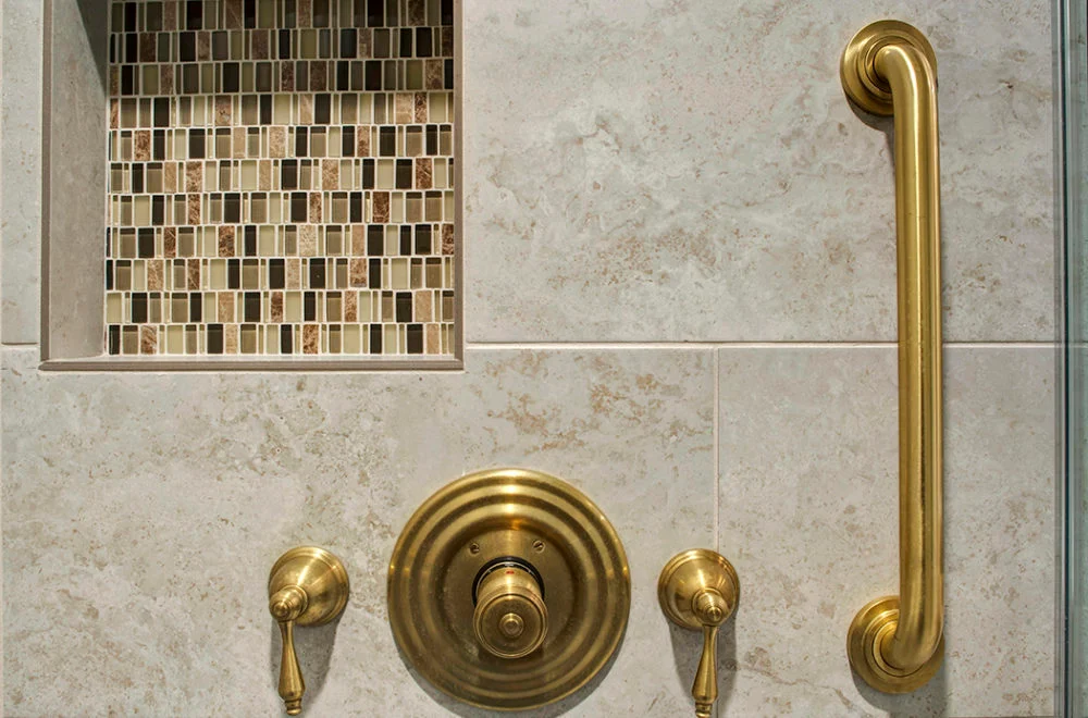 Close-up of a shower wall with beige tiles, a gold grab bar, two gold handles, a gold shower control, and a recessed shelf with mosaic tiles.