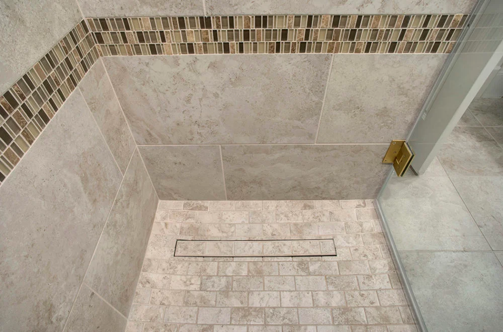 A tiled shower floor with a linear drain, beige tiles, a strip of mosaic tile accent, and a glass shower door with a metal hinge.