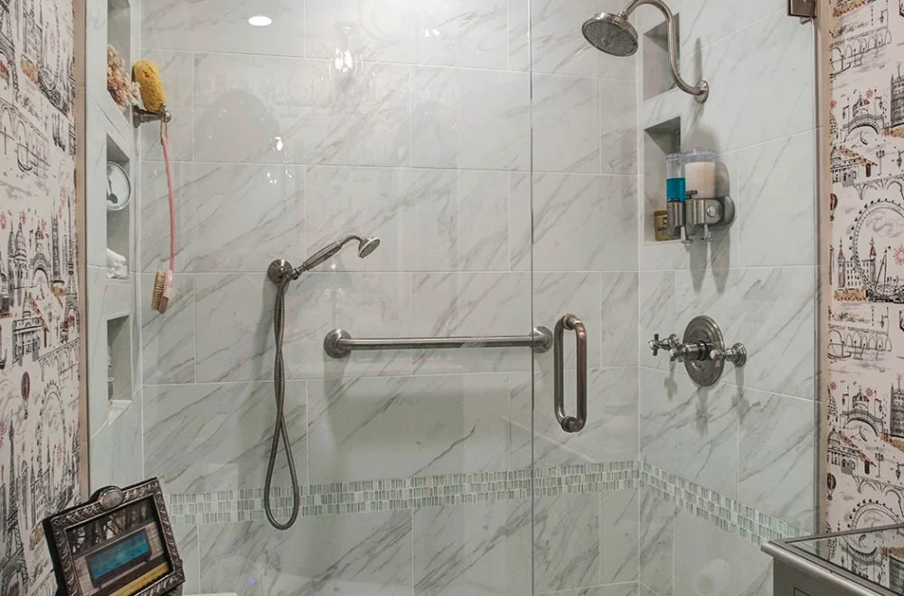 Glass-enclosed shower with marble-patterned tiles, a wall-mounted showerhead, handheld shower, grab bar, and built-in shelf holding toiletries.