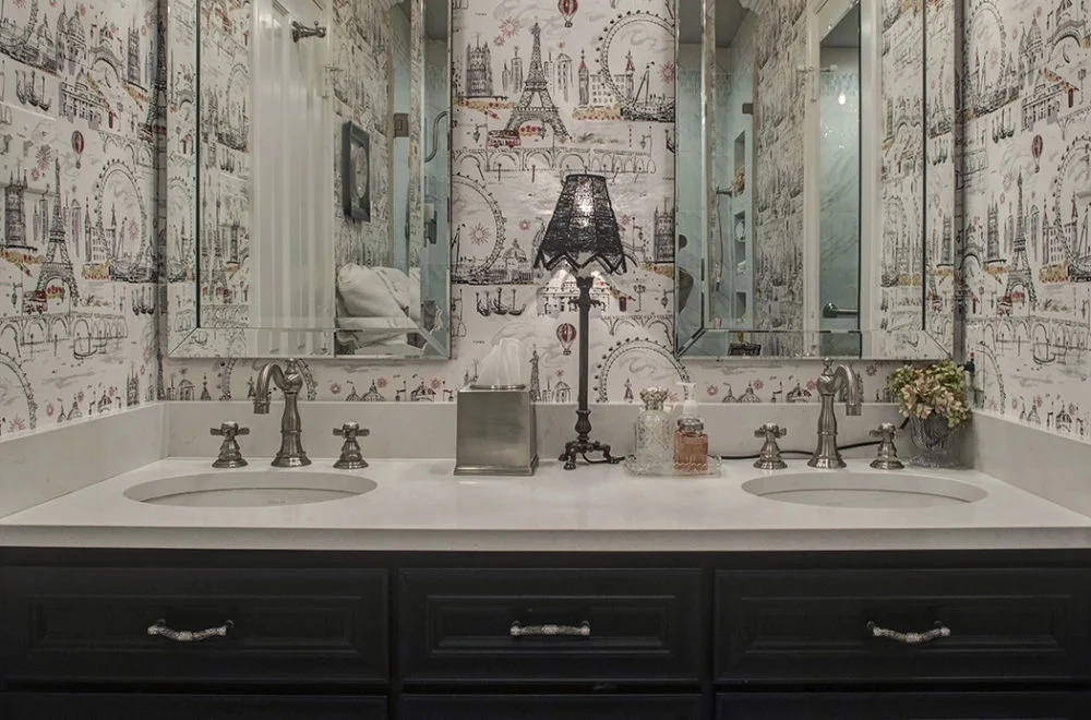 Double sink bathroom vanity with dark cabinets, two mirrors, patterned wallpaper, and various countertop items including a lamp, tissue box, and small decorative accessories.