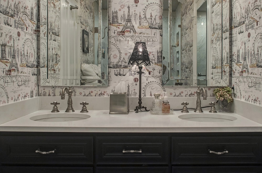 A bathroom with a double sink vanity, dark cabinets, two mirrors, a tissue box, soap dispensers, a small lamp, and decorative wallpaper featuring cityscapes.