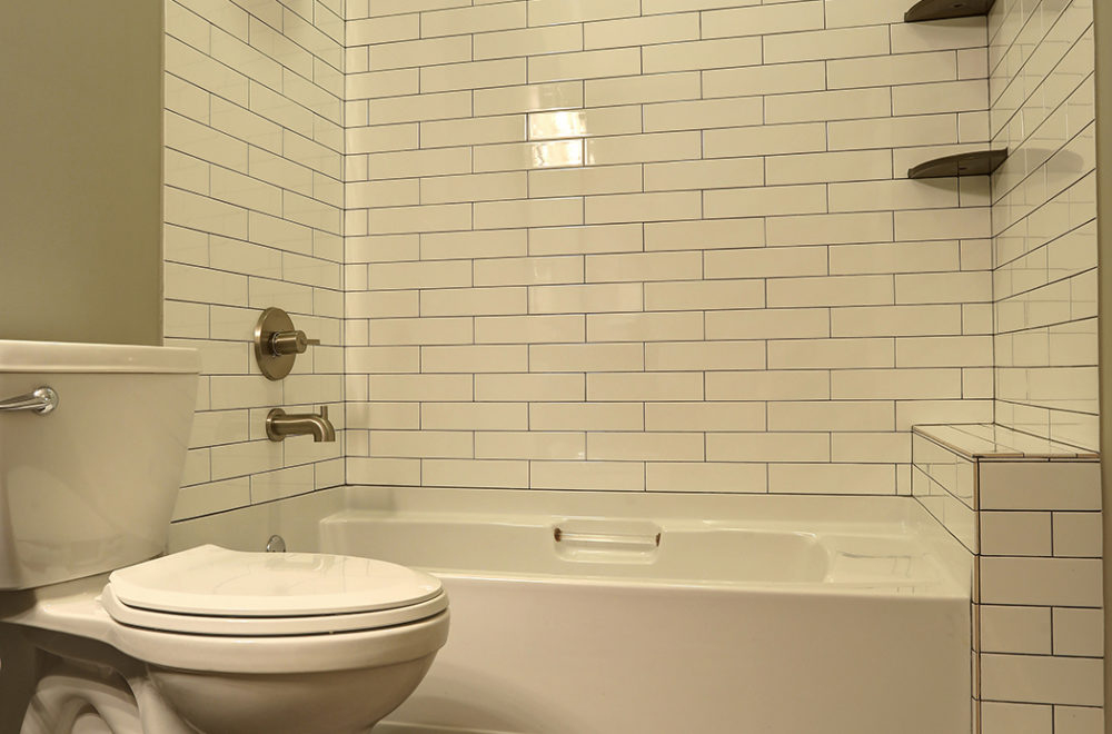 A bathroom with a white toilet, a bathtub with built-in shelving, and white subway tile walls with light grout.
