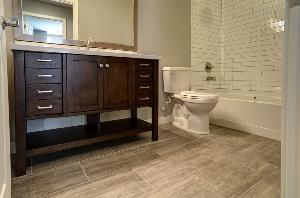 Modern bathroom with wood vanity, white toilet, and bathtub with white subway tile surround; light brown tile flooring and neutral walls.