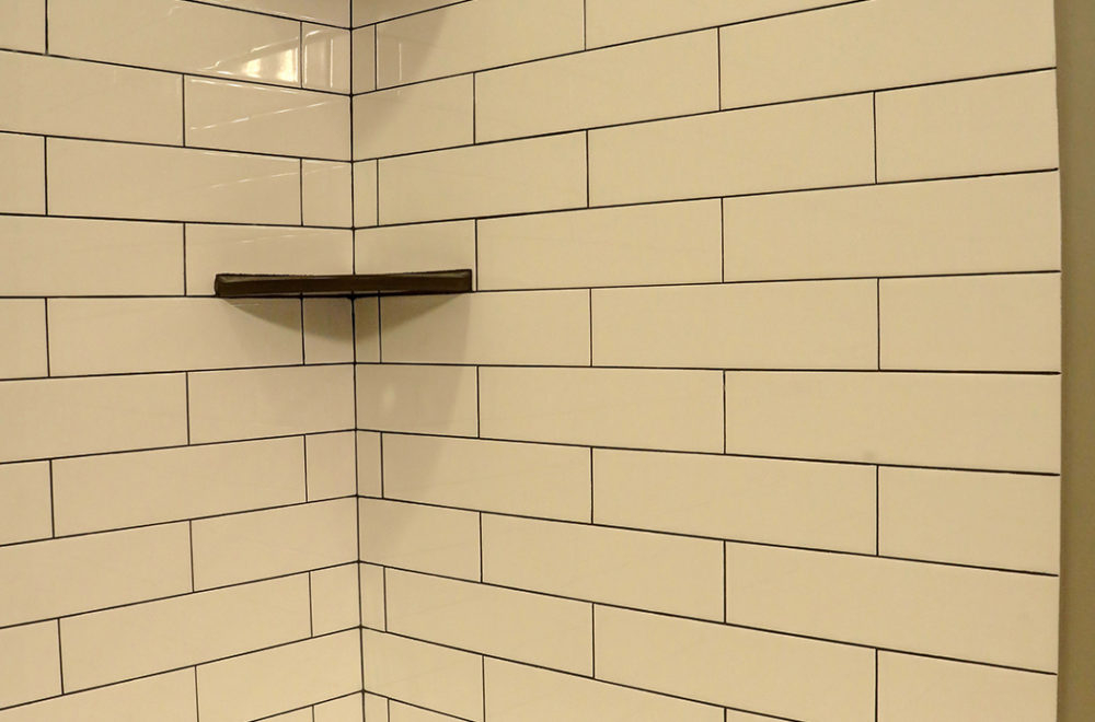 White subway tiles with dark grout cover the corner walls of a shower, featuring a small dark wooden shelf mounted on one side.