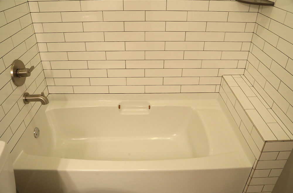 A white bathtub with a tiled surround and built-in ledge, featuring stainless steel faucet and handle fixtures, against white subway tile walls.