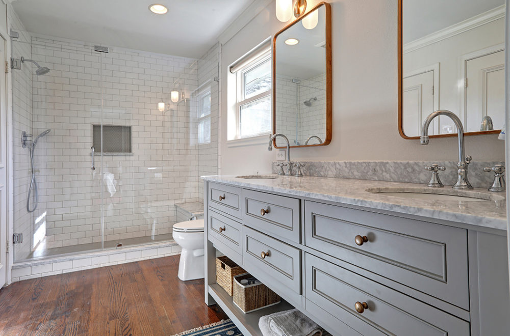 Modern bathroom with double-sink vanity, marble countertop, large mirrors, wood flooring, glass-enclosed shower, and white subway tile walls.