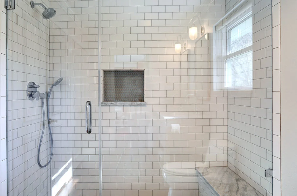 Modern shower with white subway tiles, a glass door, wall-mounted showerhead, built-in niche, marble bench, and natural light from a nearby window.
