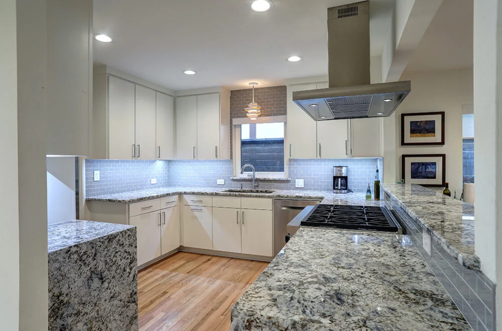 Modern kitchen with granite countertops, white cabinets, stainless steel appliances, under-cabinet lighting, and wood flooring. A window is above the sink, and framed art hangs on the wall.