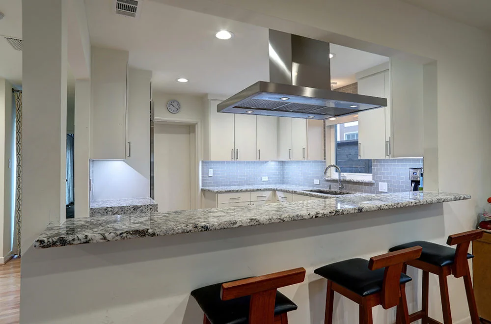 Modern kitchen with granite countertop, bar stools, stainless steel appliances, white cabinets, and under-cabinet lighting.