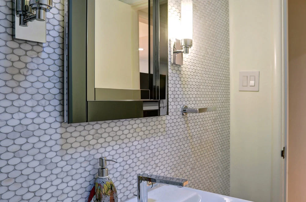 Close-up of a modern bathroom with a round tile backsplash, rectangular mirror, wall-mounted light fixture, and a sink with a soap dispenser.