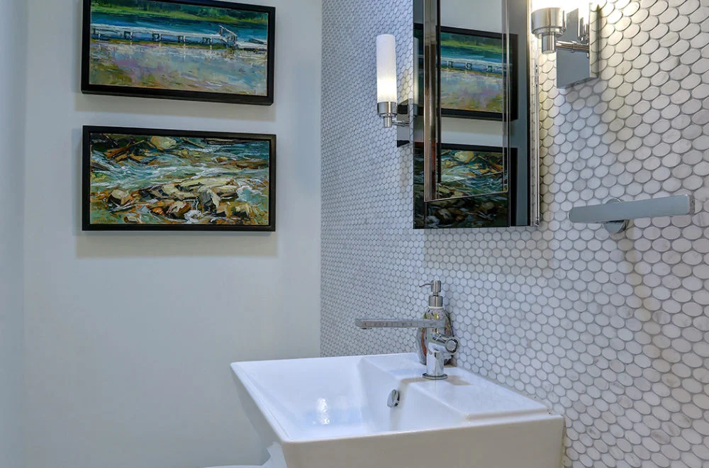 Modern bathroom with white penny tile walls, a rectangular sink, wall-mounted faucet, two landscape paintings, and a wall sconce next to a mirrored cabinet.