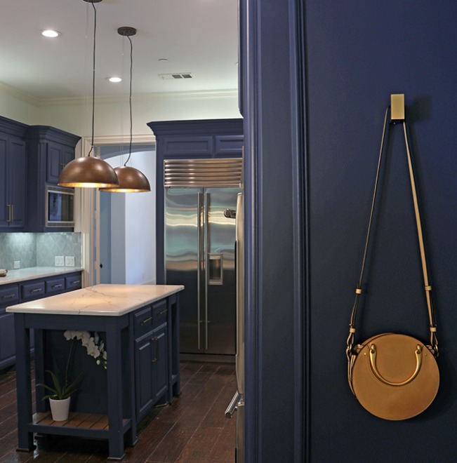 A modern kitchen with blue cabinets, a center island, stainless steel appliances, brass pendant lights, and a round tan purse hanging on a hook on the wall.