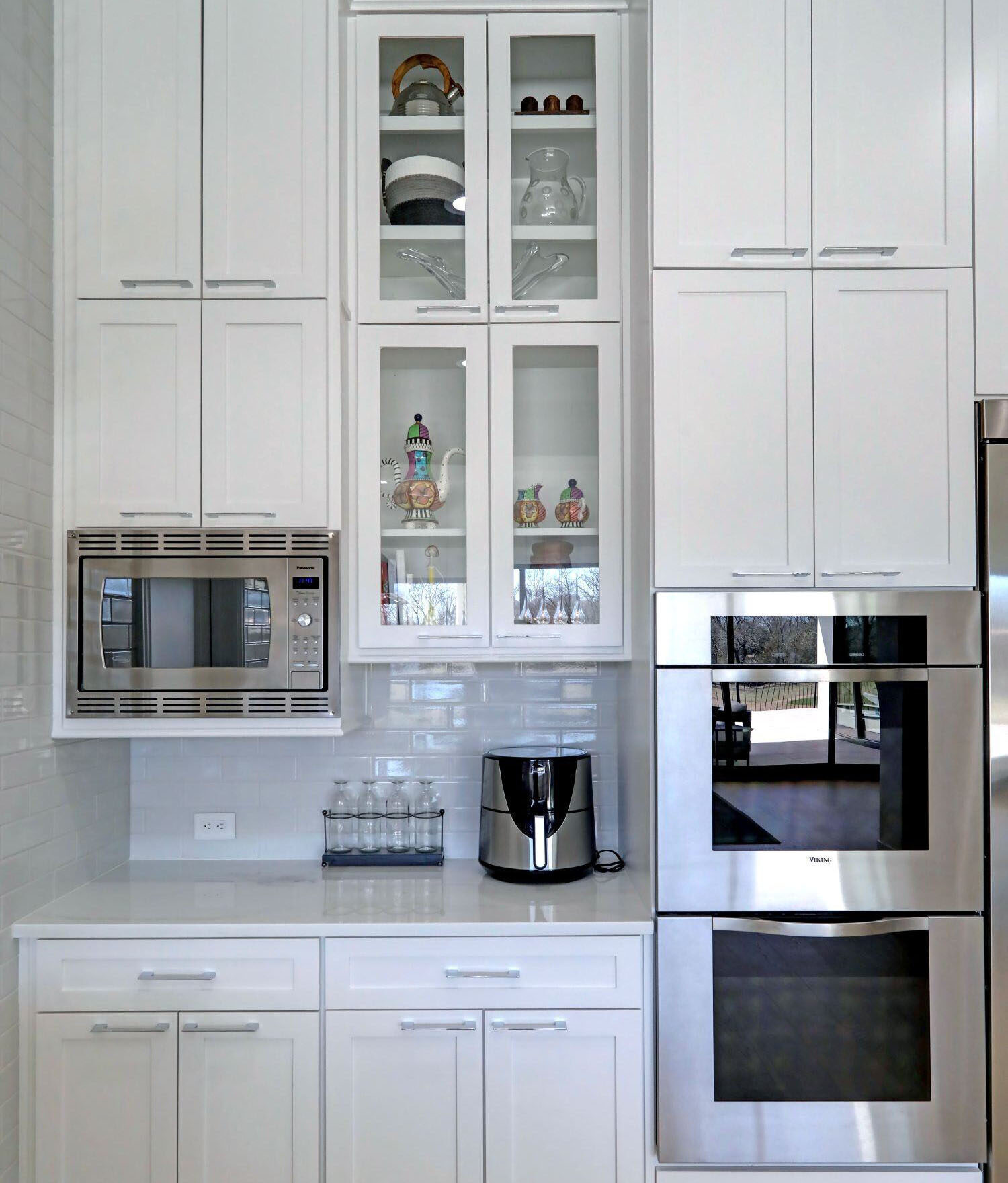 Modern white kitchen with built-in microwave, double ovens, glass-front cabinets displaying dishes, an air fryer, and a set of glasses on the countertop.