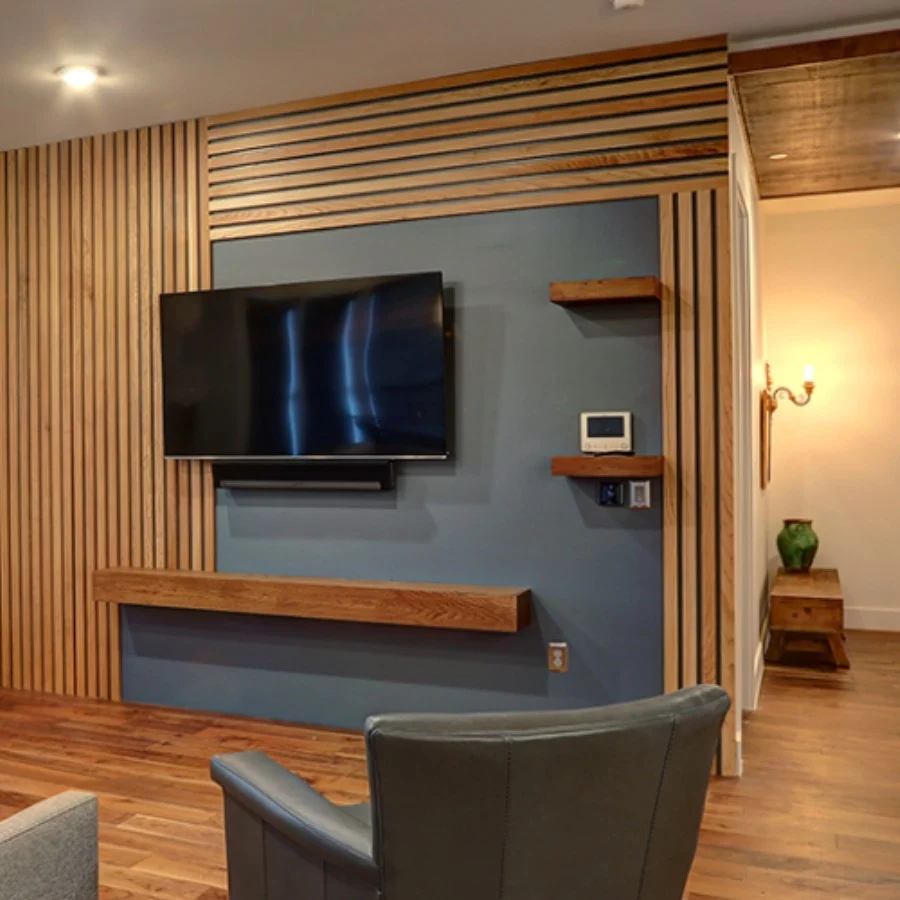Transom-Banners1 A modern living room wall with wooden slats, a mounted flat-screen TV, floating shelves, and a gray accent area with a thermostat and control panel.