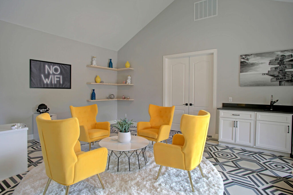 A modern lounge with four yellow armchairs around a round table, geometric floor tiles, wall shelves with decor, and a "No WiFi" sign on the wall.