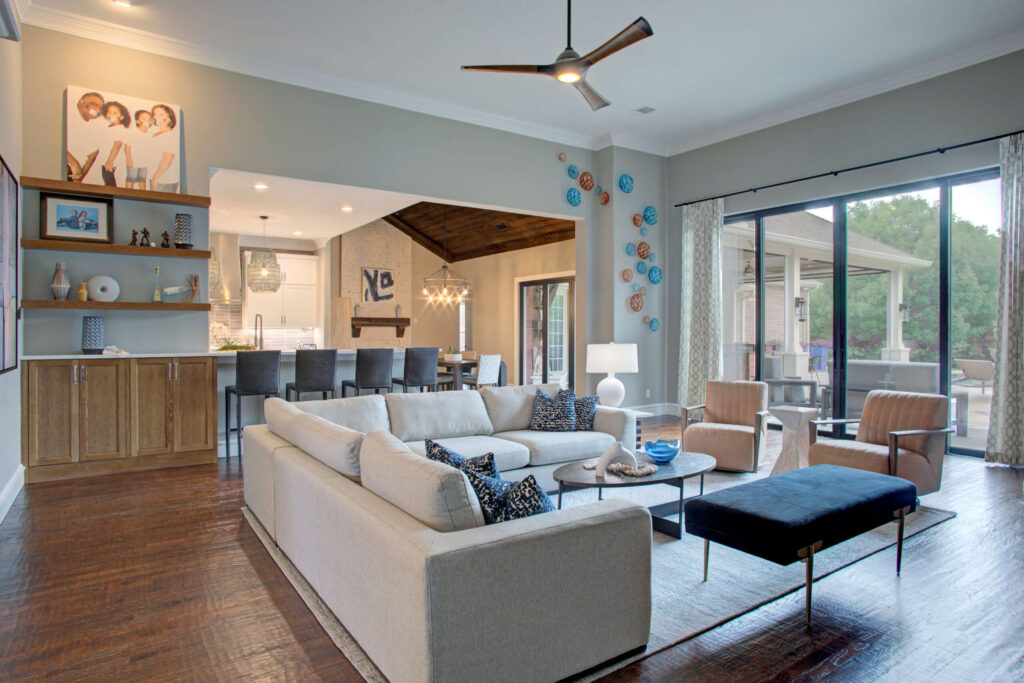 Modern open-concept living room with a large sectional sofa, two armchairs, wall art, and large windows leading to an outdoor patio. The space connects to a kitchen and dining area.