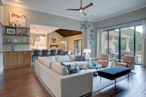 Modern open-concept living room with a large sectional sofa, two armchairs, wall art, and large windows leading to an outdoor patio. The space connects to a kitchen and dining area.