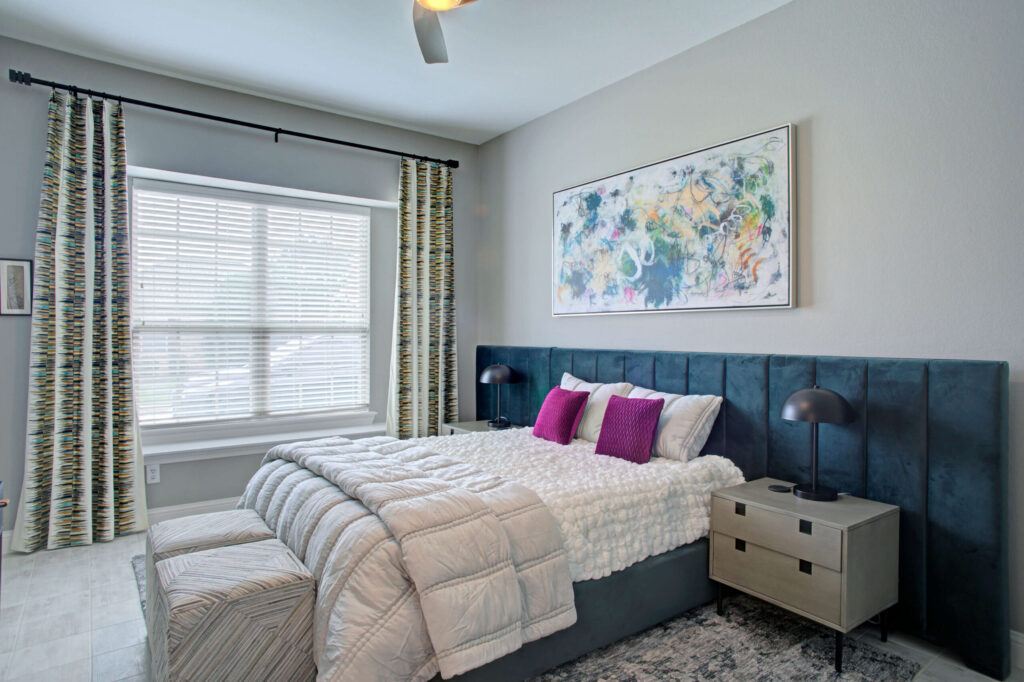Modern bedroom with a large bed, white bedding, two magenta pillows, two nightstands with lamps, patterned curtains, and abstract art on the wall.