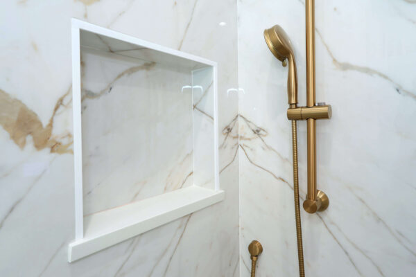 Marble shower wall with a built-in white niche and a gold handheld showerhead and fixtures.
