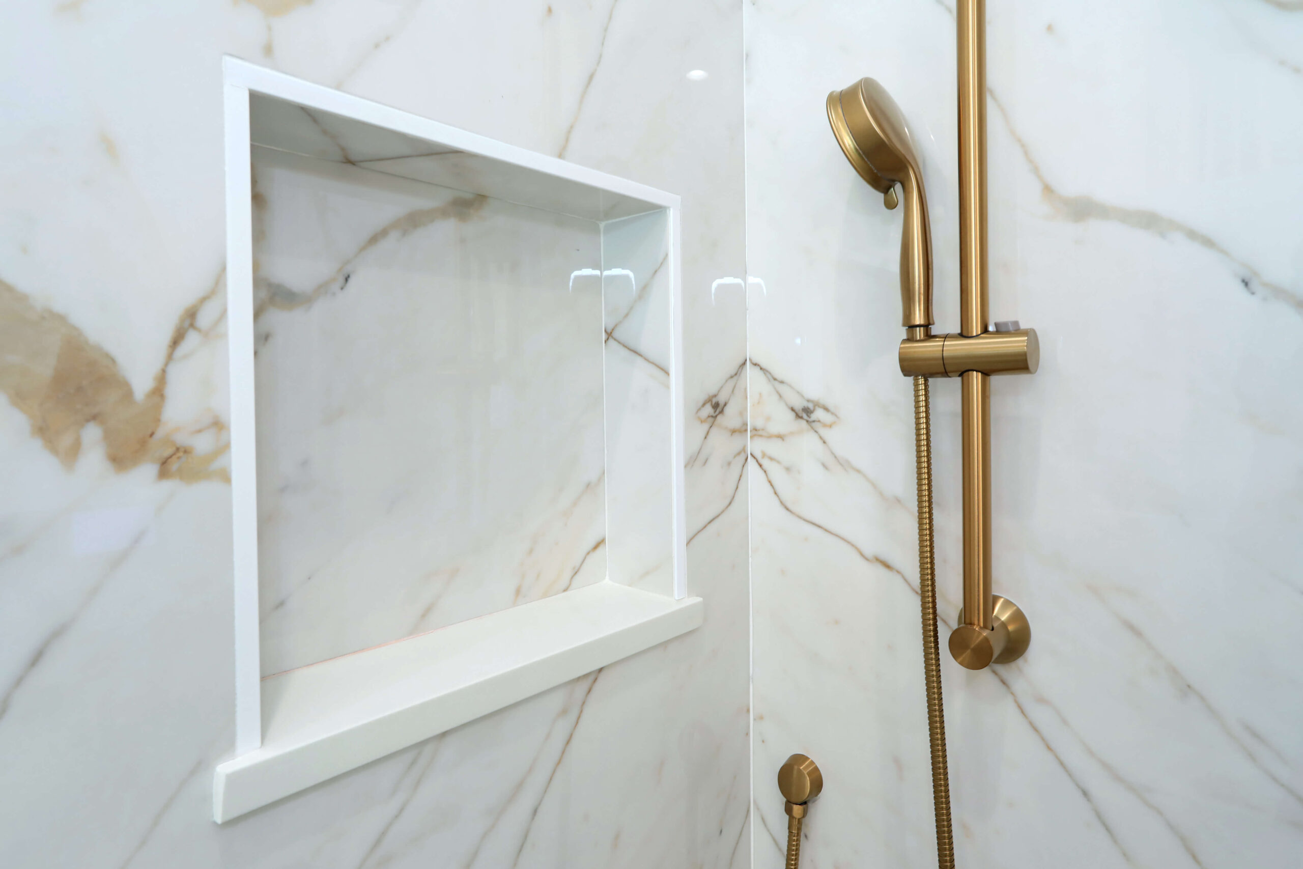 Marble shower wall with a built-in white niche and a gold handheld showerhead and fixtures.