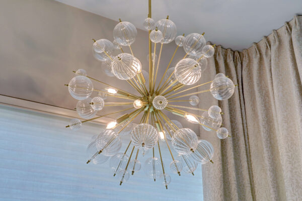 A modern chandelier with a gold frame and multiple glass globes, hanging from the ceiling near beige curtains and a large window.