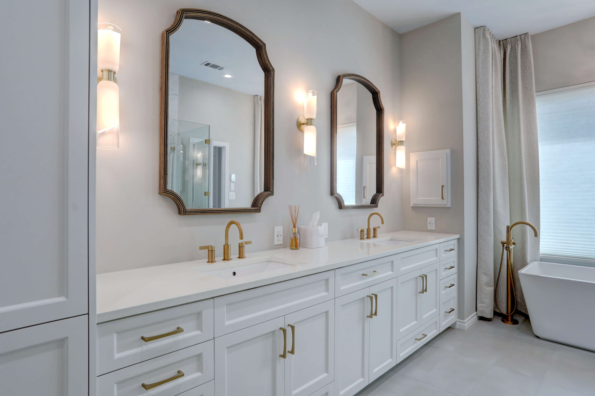 Modern bathroom with double vanity, white cabinets, brass hardware, two mirrors, twin wall sconces, and a freestanding bathtub near a large window with curtains.