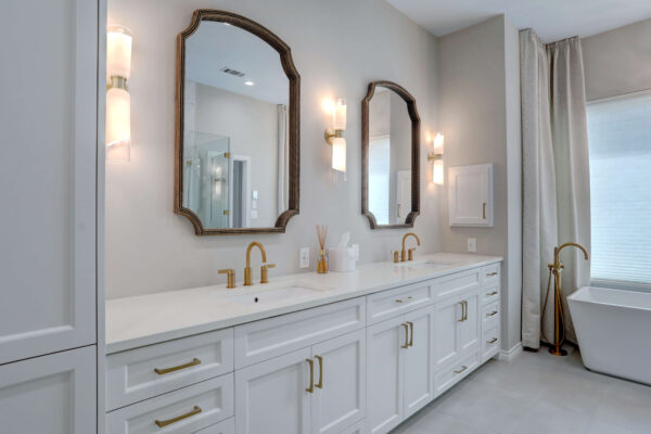 Modern bathroom with double vanity, white cabinets, brass hardware, two mirrors, twin wall sconces, and a freestanding bathtub near a large window with curtains.