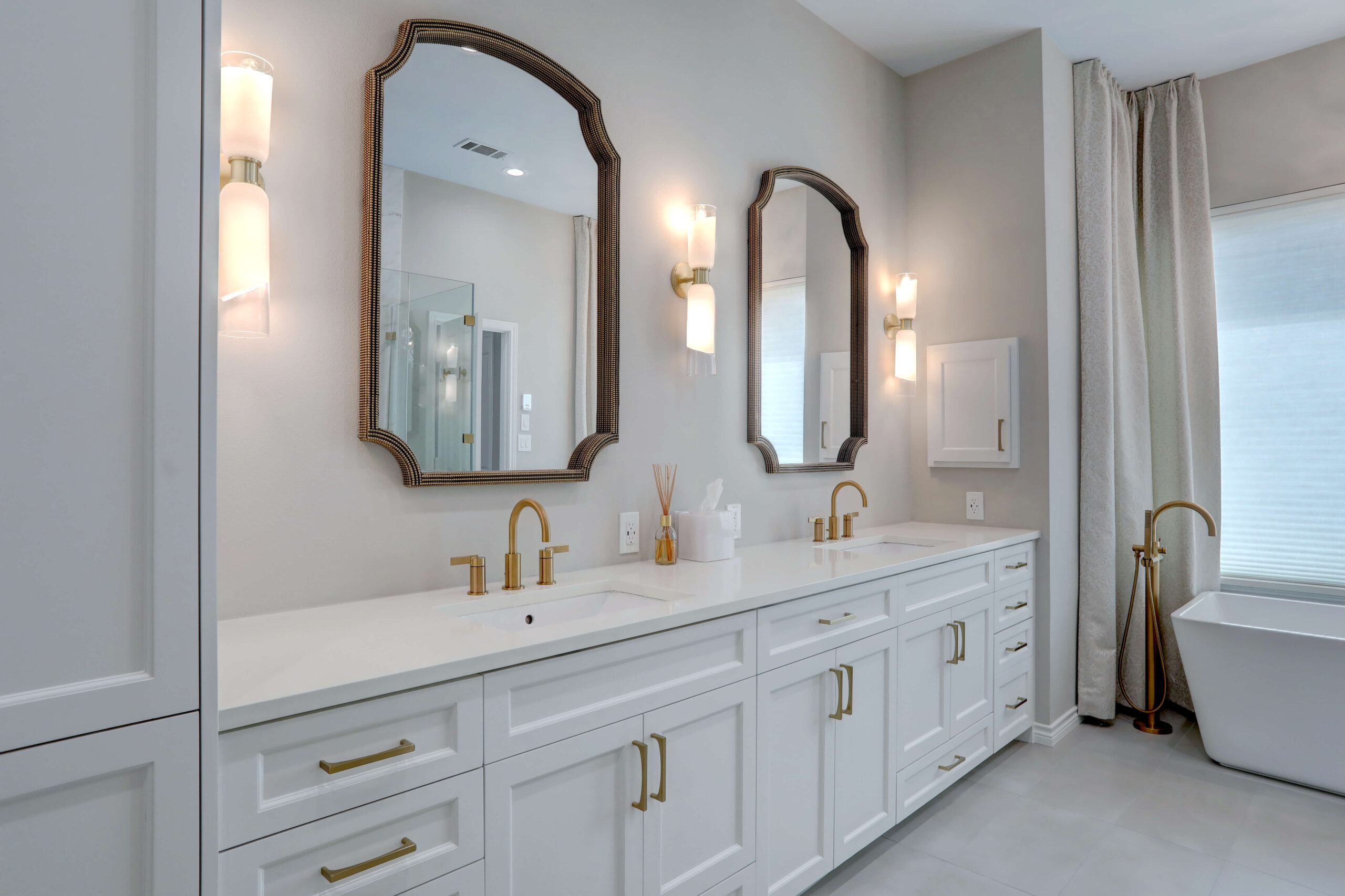 Modern bathroom with double vanity, white cabinets, brass hardware, two mirrors, twin wall sconces, and a freestanding bathtub near a large window with curtains.