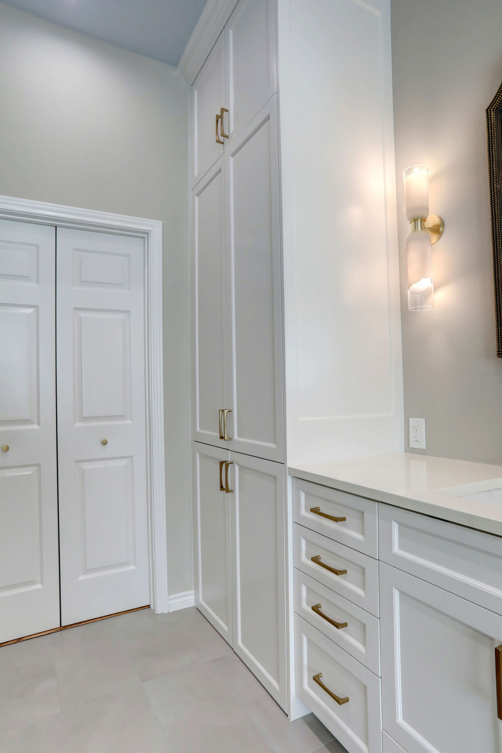 White built-in cabinets with gold handles next to a double door closet, under a wall-mounted light fixture, in a modern interior space.