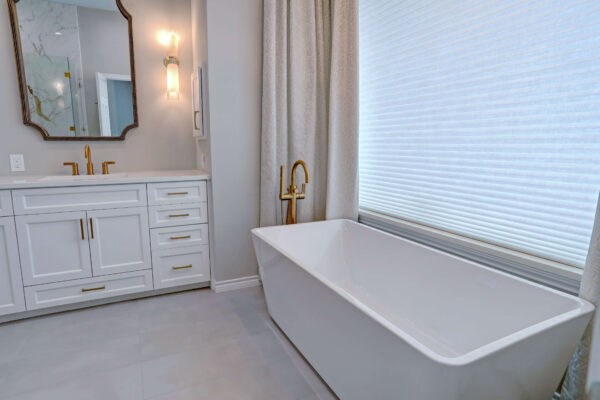 Modern bathroom with a white freestanding bathtub, gold faucet, white vanity with gold handles, large mirror, and window with blinds.