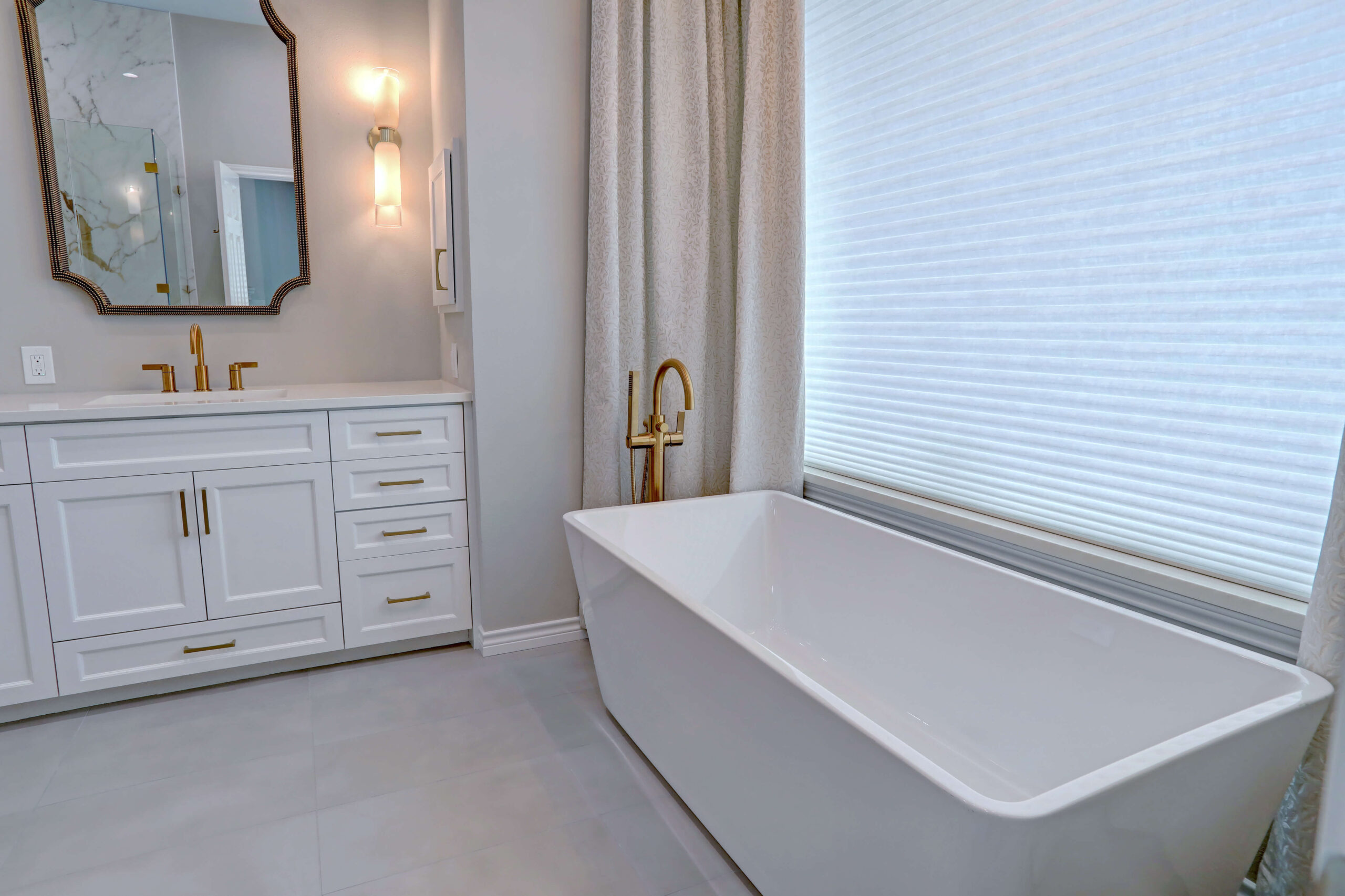 Modern bathroom with a white freestanding bathtub, gold faucet, white vanity with gold handles, large mirror, and window with blinds.
