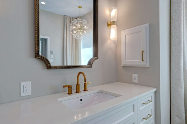 Modern bathroom vanity with a white countertop, gold faucet, mounted mirror, wall sconce, and a small white cabinet on a light gray wall.
