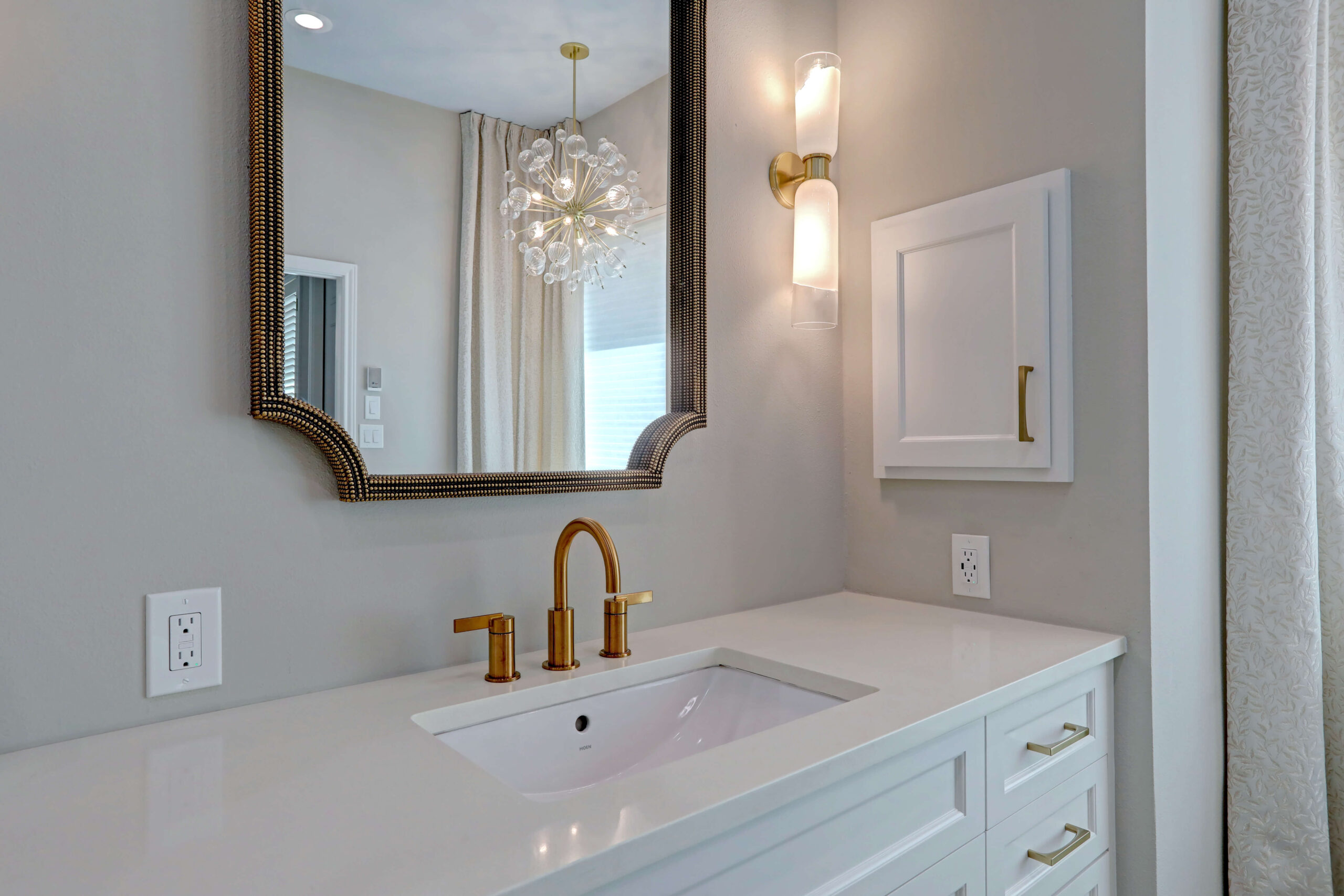 Modern bathroom vanity with a white countertop, gold faucet, mounted mirror, wall sconce, and a small white cabinet on a light gray wall.
