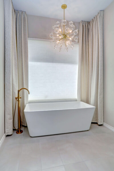 A modern bathroom features a white rectangular freestanding tub, gold floor-mounted faucet, white drapes, and a contemporary chandelier.