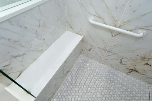 Marble shower with a built-in white bench, a safety grab bar on the wall, and a floor with small hexagonal tiles.