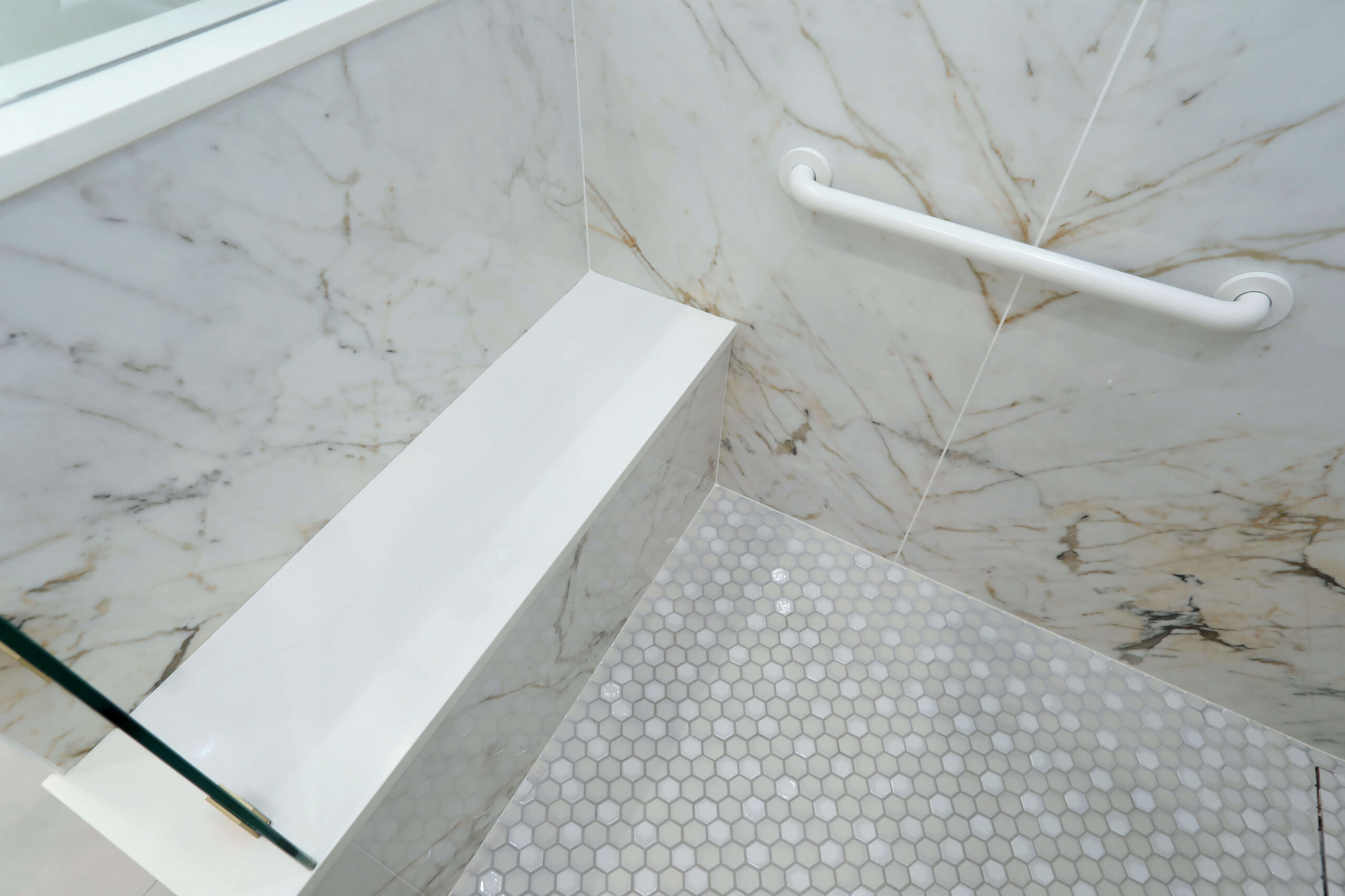 Marble shower with a built-in white bench, a safety grab bar on the wall, and a floor with small hexagonal tiles.
