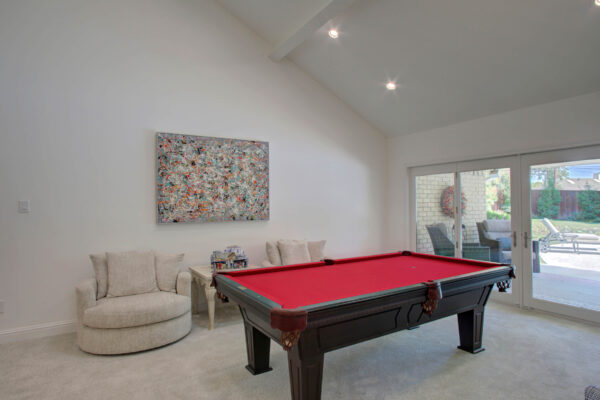 Perfect for planning a home addition, this room features a red-felt pool table, two beige armchairs, abstract wall art, and sliding glass doors leading to an outdoor patio.