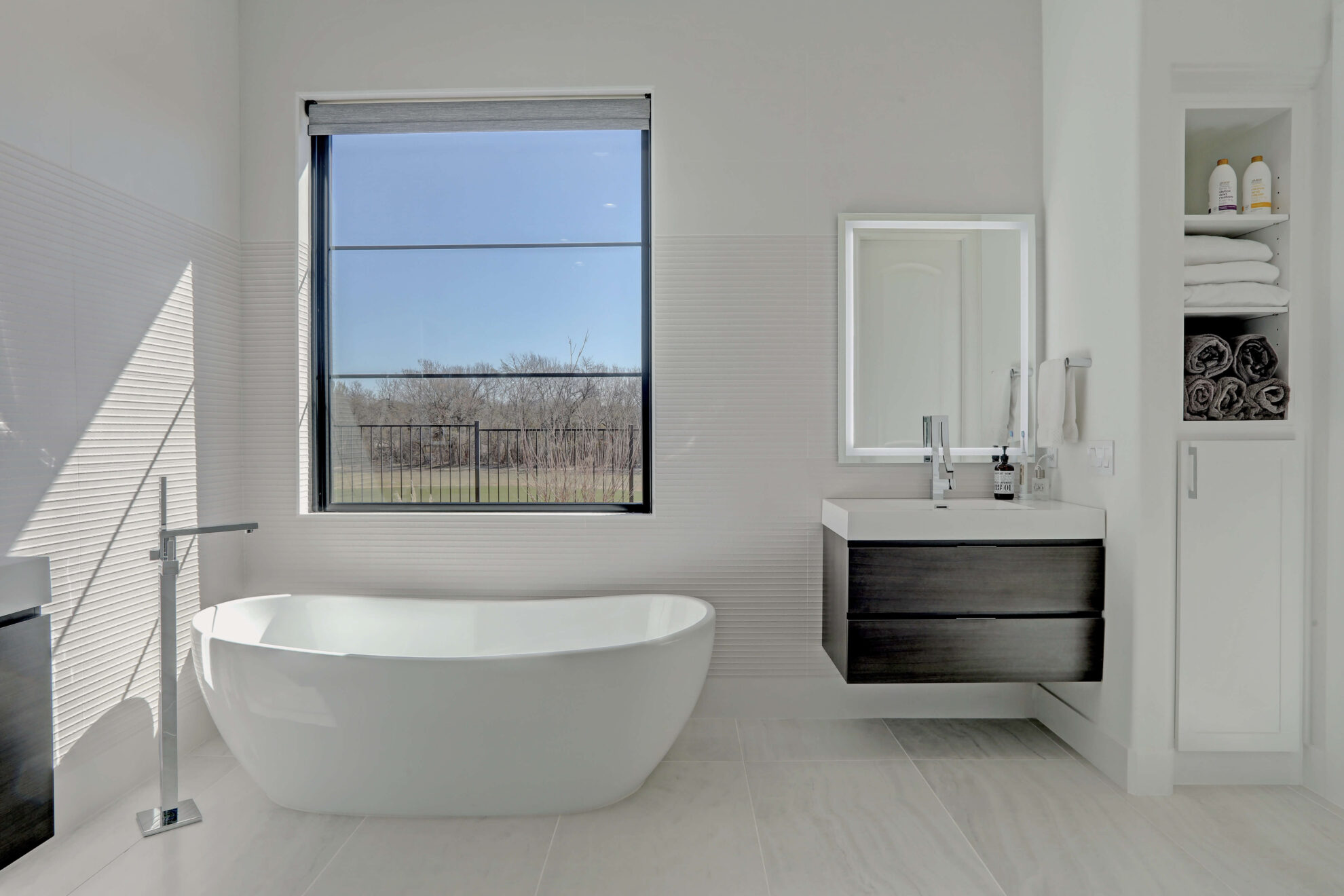 Modern bathroom with a freestanding bathtub, large window, wall-mounted sink cabinet, shelves with towels and bath products—perfect inspiration if you’re planning a home addition filled with natural light.