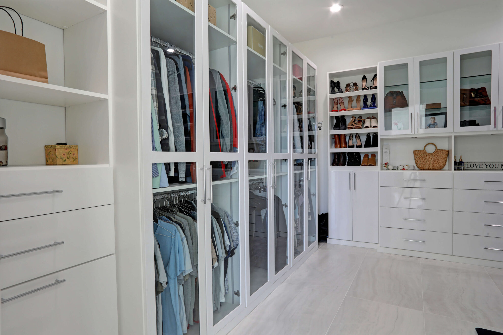 A modern walk-in closet with white cabinetry, glass-front wardrobes displaying clothes, shelves with shoes and bags, and drawers for storage—an ideal feature to consider when planning a home addition.