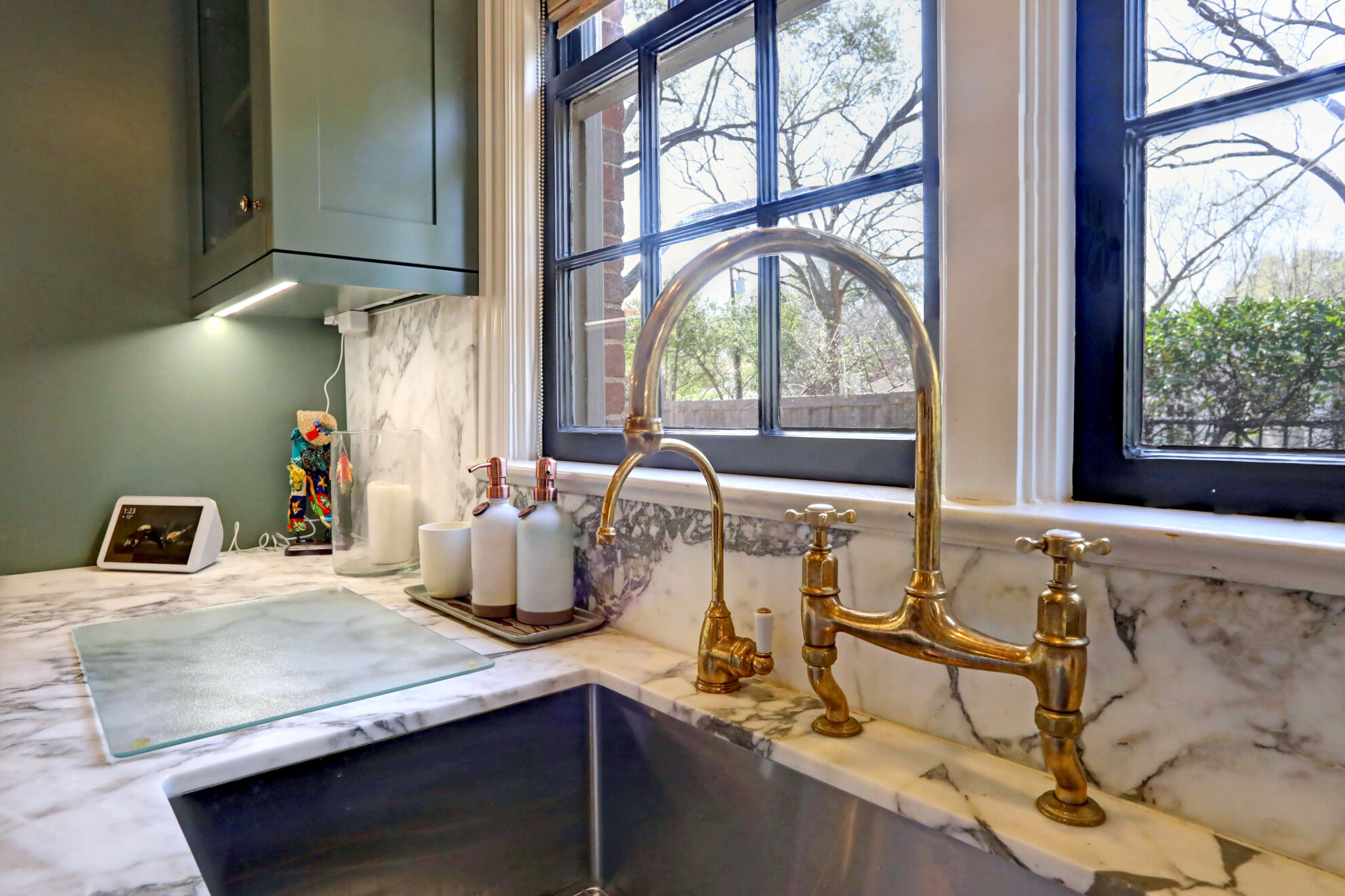A brass kitchen faucet is installed over a marble countertop and sink, with soap dispensers and a cutting board nearby—perfect inspiration if you’re planning a home addition that highlights natural light and elegant finishes.