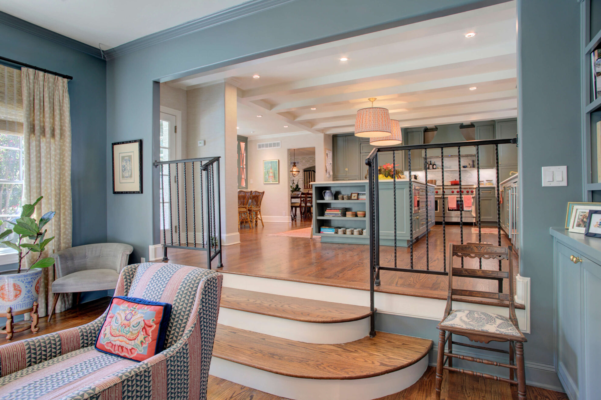 Open-concept living space with blue walls, wood floors, a raised kitchen area, steps with black railings, and modern furnishings—perfect inspiration if you’re planning a home addition.