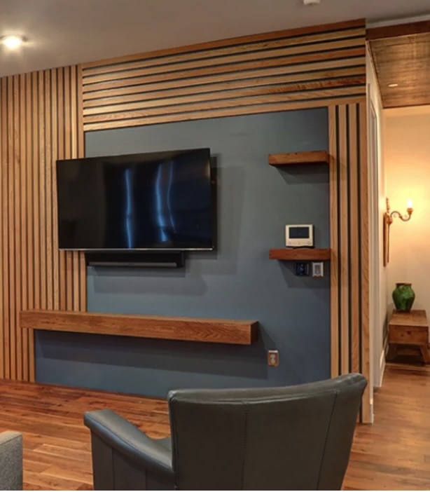A modern living room wall with wood slats, a mounted flat-screen TV, floating shelves, and a gray-blue accent background. A gray chair is partially visible in the foreground.