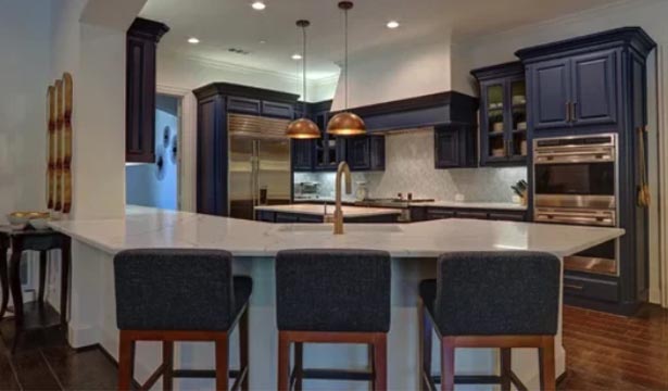 Modern kitchen with dark blue cabinets, stainless steel appliances, a large white marble island, gold faucet, and three dark upholstered barstools.