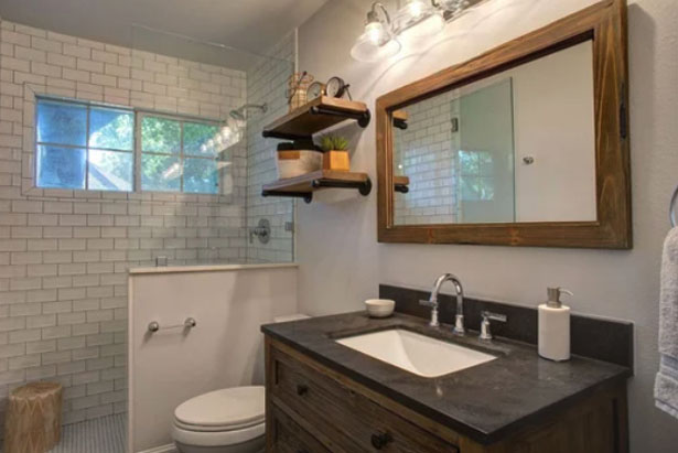 Modern bathroom with a wooden vanity, rectangular mirror, open shelves with decor, white subway tile walls, a toilet, and a glass-enclosed shower with a window.