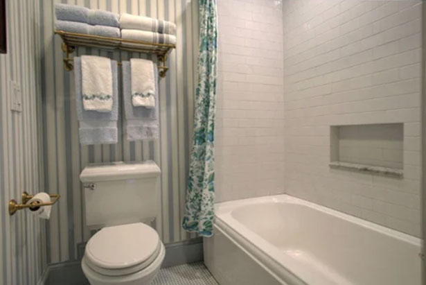 A bathroom with a toilet, bathtub with a floral shower curtain, white subway tile walls, and shelves above the toilet holding towels.
