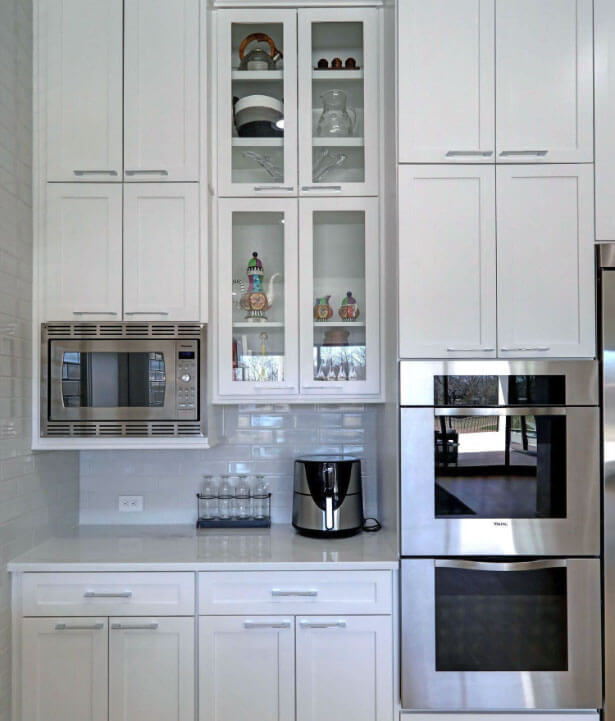 Modern kitchen with white cabinets, built-in microwave, double ovens, glass display shelves, countertop appliance, and a row of glass jars on the counter.